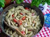 En stor panna med krämig pasta pesto, körsbärstomater och kyckling. En träsked ligger i pannan.