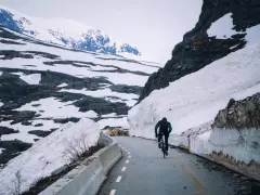 En person cyklar på en snöig väg i Norge, omgiven av höga snödrivor och berg.