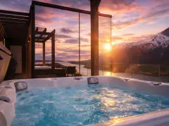 Bubblande badtunna med utsikt över en fjord och snöklädda berg i Innerdalen, Norge. Solnedgången färgar himlen i varma toner.