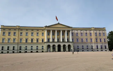 Det gula **Oslo slott** med sin flagga reser sig stolt mot en klarblå himmel. En stor öppen plats framför slottet.