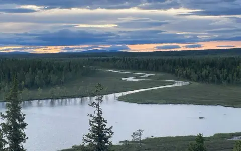 En slingrande flod genom ett vidsträckt skoglandskap i Muddus nationalpark. Molnen på himlen färgas av solnedgången.