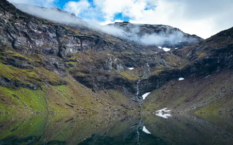 En spegelblank sjö reflekterar en dramatisk bergssida med en forsande vattenfall. Perfekt för att vandra i Abisko.