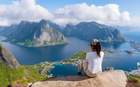 En person sitter på en klippa och blickar ut över en dramatisk kustlinje med berg och fjordar. En perfekt vy för lofoten vandring.