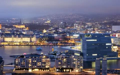 Fakta om Oslo: En stad vid vattnet med moderna byggnader och upplysta hus, som sträcker sig mot en kulle i fjärran.