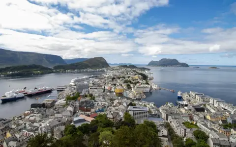 Vacker vy över Ålesund, Norge, med färgglada hus, fartyg i hamnen och berg i bakgrunden under en molnig himmel.
