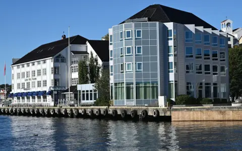 Hotellbygg vid vattnet i Arendal, Norge. En vit byggnad med blå markiser och en modern glasbyggnad står sida vid sida.