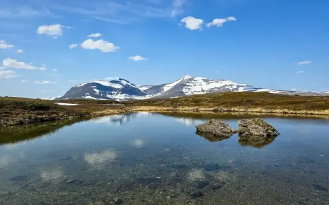 En klarblå himmel med moln speglar sig i en lugn sjö. I bakgrunden reser sig snöklädda berg, en vacker vy för ramundberget vandring.