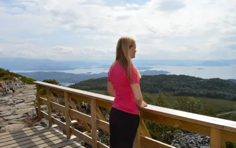 Kvinna i rosa tröja njuter av utsikten över ett storslaget Norge med fjäll och fjordar.