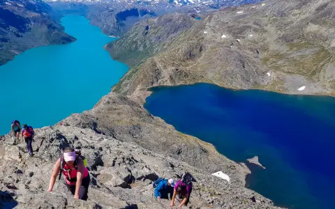 Vandrare klättrar på en stenig bergssida med turkosa och blå sjöar nedanför. En klassisk Besseggen vandring.