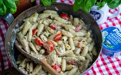 En stor panna med krämig pasta pesto, körsbärstomater och kyckling. En träsked ligger i pannan.