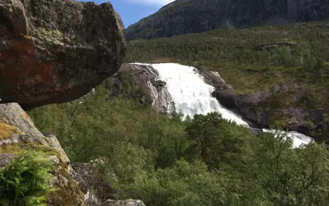 En mäktig fors forsar nerför bergssluttningen, omgiven av grönskande skog och stenblock. En perfekt vy för vandring på Hardangervidda.