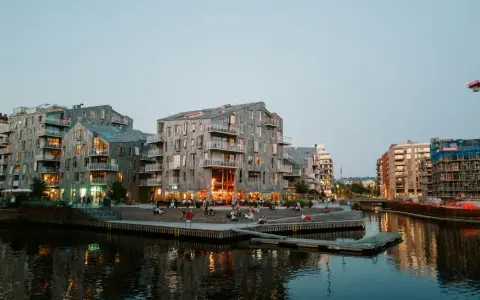 Moderna byggnader längs en kanal i Oslo. Folk sitter på trappor vid vattnet. Besök Oslo för en härlig kväll.