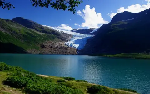 Blå sjö vid foten av en glaciär i Steinkjer, Norge. Grönskande bergssluttningar möter det turkosa vattnet under en klarblå himmel.