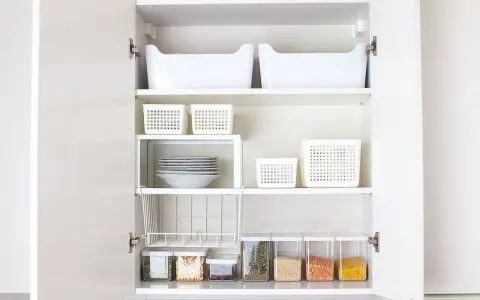 Vitt skåp med hyllor fyllda med vita förvaringskorgar, staplade tallrikar och genomskinliga burkar med matvaror. En perfekt organiserad hylla i vardagsrummet.