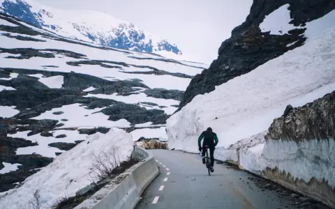 En person cyklar på en snöig väg i Norge, omgiven av höga snödrivor och berg.