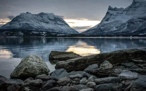 Vinterlandskap med snöklädda berg i Lyngenalperna, speglade i ett lugnt vatten. Stenar ligger vid strandkanten.