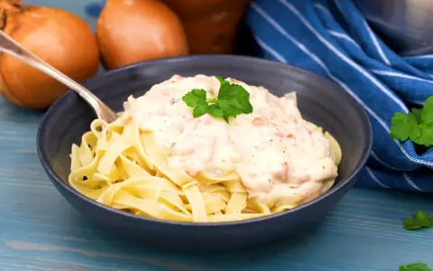 En skål med tagliatelle täckt av krämig ost och skinksås, garnerad med persilja. Två gula lökar syns i bakgrunden.