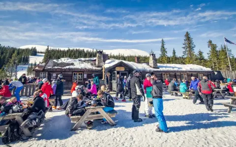 Folk njuter av afterski i Trysil vid en mysig timmerstuga med snöklädda berg och granar i bakgrunden.