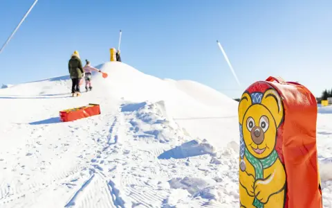 En glad björn pryder en pulka backe. Barn klädda för kyla väntar på sin tur att åka nerför den snöiga kullen.