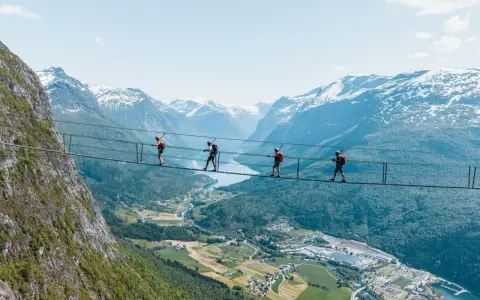 Fyra personer går på en hängbro vid Loen via ferrata, med en fantastisk utsikt över fjord och snöklädda berg.