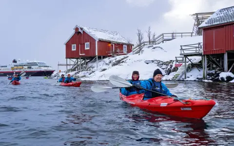 Glada paddlare i kajak i Lofoten, med snöklädda röda hus och ett stort fartyg i bakgrunden.