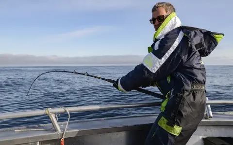 Man med solglasögon och fiskeutrustning kämpar med en böjd spö ombord på en båt. En spännande dag med fiska i Norge.