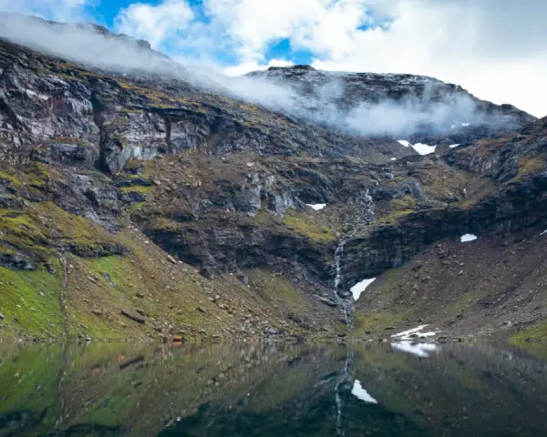 En spegelblank sjö reflekterar en dramatisk bergssida med en forsande vattenfall. Perfekt för att vandra i Abisko.