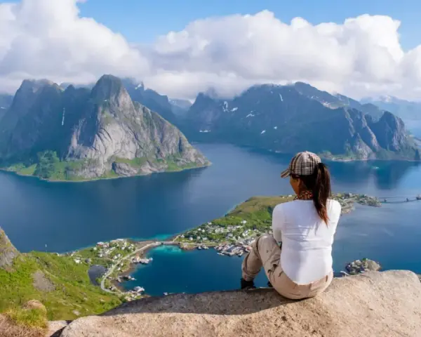 En person sitter på en klippa och blickar ut över en dramatisk kustlinje med berg och fjordar. En perfekt vy för lofoten vandring.