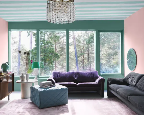 Ett vardagsrum med art deco inredning, mjuklila soffa, mörkgrå soffa, turkos pall och en stor fönsterutsikt.