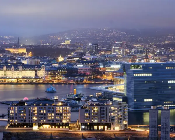 Fakta om Oslo: En stad vid vattnet med moderna byggnader och upplysta hus, som sträcker sig mot en kulle i fjärran.