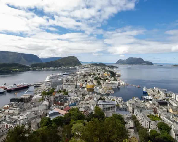 Vacker vy över Ålesund, Norge, med färgglada hus, fartyg i hamnen och berg i bakgrunden under en molnig himmel.