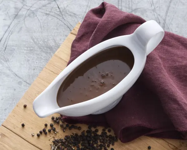 En vit såsslev fylld med mustig brunsås, bredvid en hög med svartpepparkorn. Ett perfekt brunsås recept för middagen.