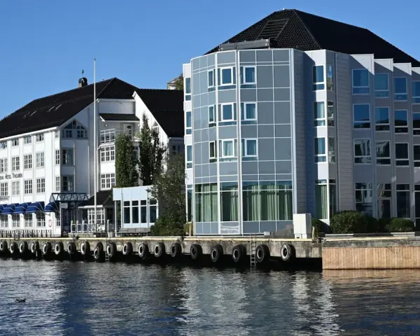 Hotellbygg vid vattnet i Arendal, Norge. En vit byggnad med blå markiser och en modern glasbyggnad står sida vid sida.