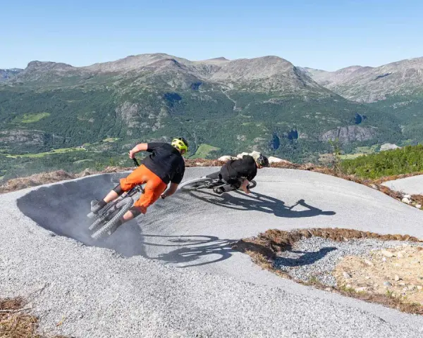 Två cyklister kör utför en grusig bana i Hemsedal sommar. Berg och grönska i bakgrunden.