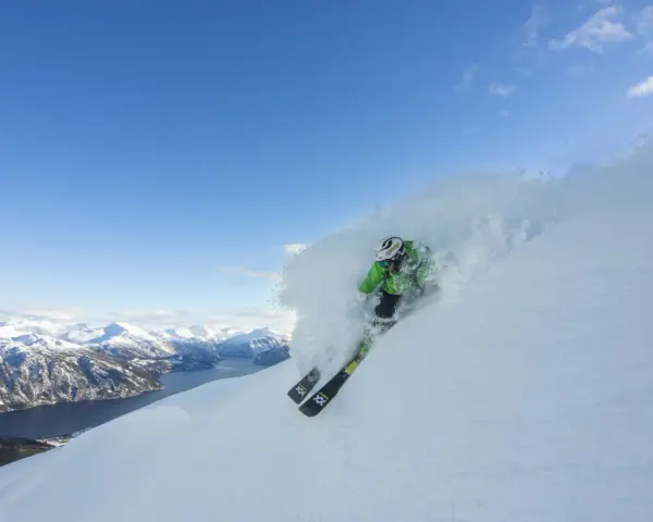 En skidåkare i grön jacka plöjer genom pudersnö med en fantastisk fjordvy i bakgrunden. Stranda ski-skidorna skär genom snön.