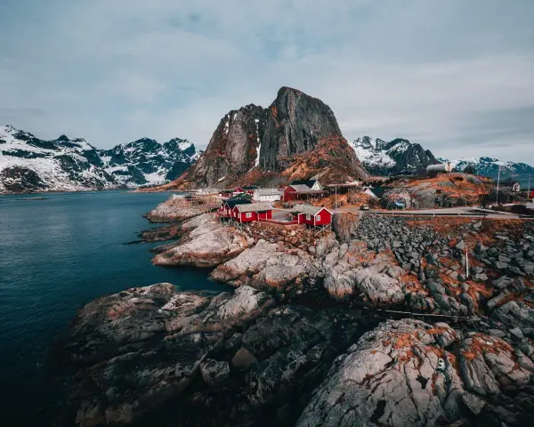 Röda fiskestugor vid en klippig kust i **Hadeland, Norge**, med snöklädda berg i bakgrunden.