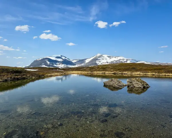 En klarblå himmel med moln speglar sig i en lugn sjö. I bakgrunden reser sig snöklädda berg, en vacker vy för ramundberget vandring.