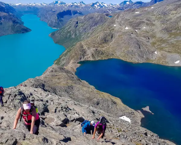 Vandrare klättrar på en stenig bergssida med turkosa och blå sjöar nedanför. En klassisk Besseggen vandring.