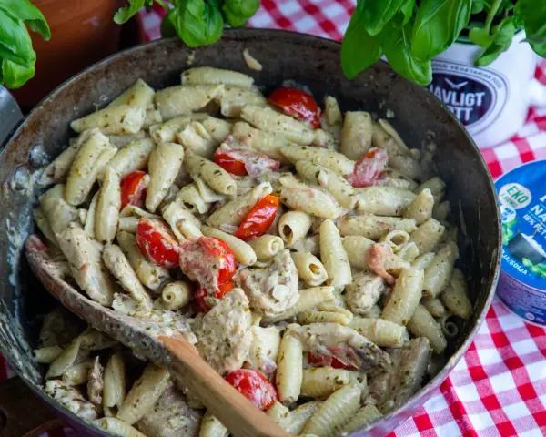En stor panna med krämig pasta pesto, körsbärstomater och kyckling. En träsked ligger i pannan.