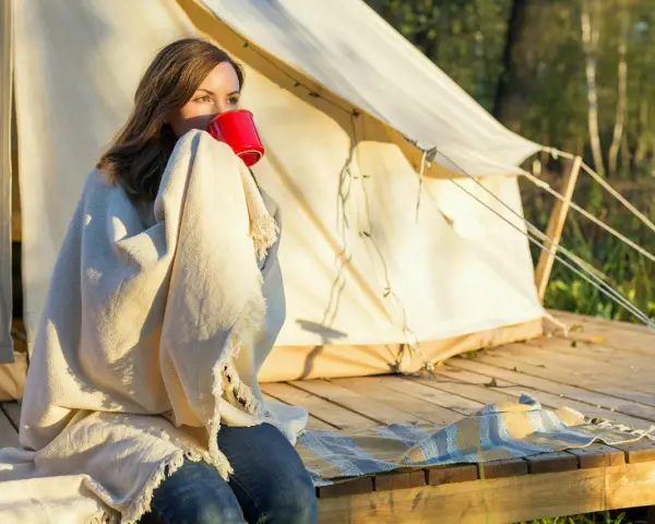 Kvinna med filt och röd mugg vid tältet. En perfekt packlista camping-morgon.