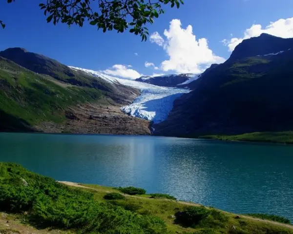 Blå sjö vid foten av en glaciär i Steinkjer, Norge. Grönskande bergssluttningar möter det turkosa vattnet under en klarblå himmel.