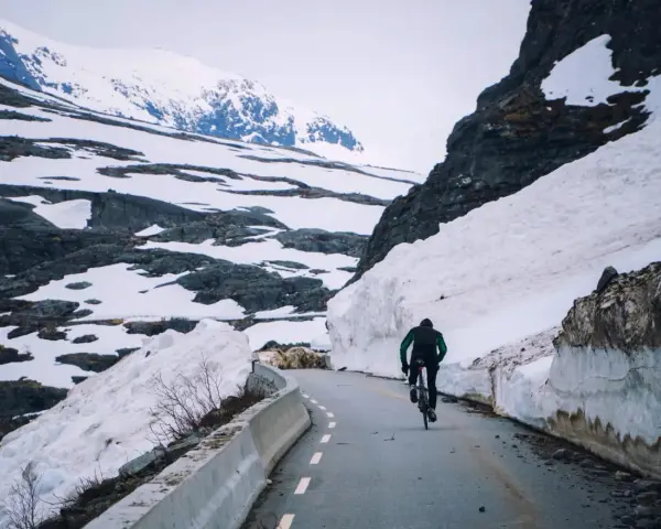 En person cyklar på en snöig väg i Norge, omgiven av höga snödrivor och berg.