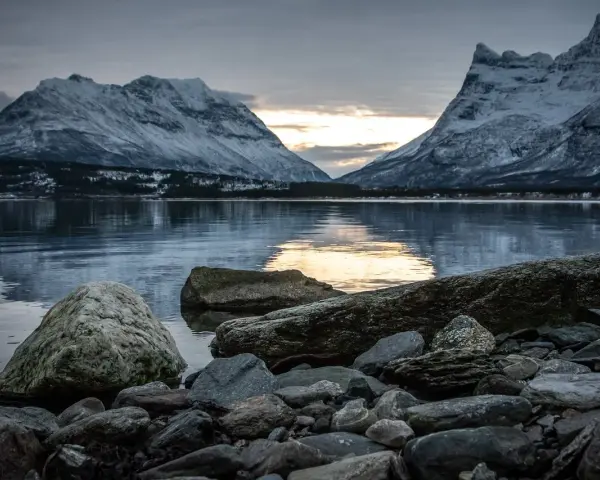 Vinterlandskap med snöklädda berg i Lyngenalperna, speglade i ett lugnt vatten. Stenar ligger vid strandkanten.