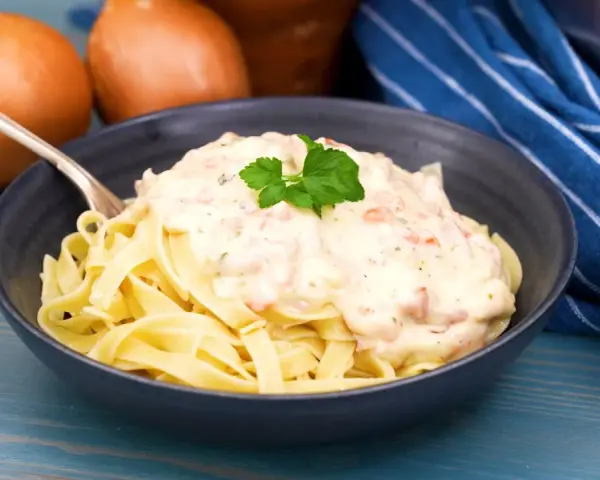 En skål med tagliatelle täckt av krämig ost och skinksås, garnerad med persilja. Två gula lökar syns i bakgrunden.