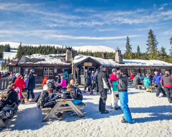 Folk njuter av afterski i Trysil vid en mysig timmerstuga med snöklädda berg och granar i bakgrunden.