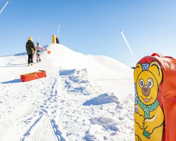 En glad björn pryder en pulka backe. Barn klädda för kyla väntar på sin tur att åka nerför den snöiga kullen.
