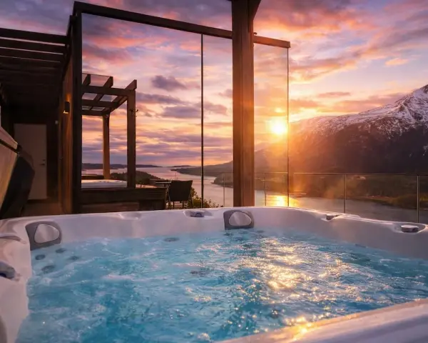 Bubblande badtunna med utsikt över en fjord och snöklädda berg i Innerdalen, Norge. Solnedgången färgar himlen i varma toner.