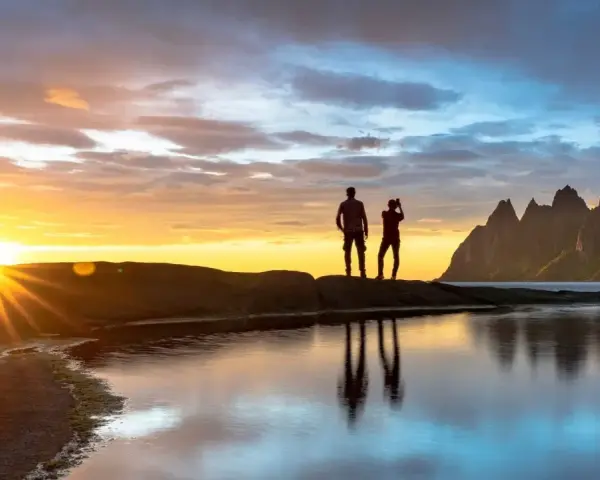 Två silhuetter står på en klippa vid havet och ser på solnedgången över Bodo, Norge. Bergskedja i bakgrunden.