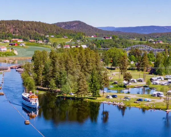 En båt på en sjö vid en campingplats i Norge. Husbilar och tält står utspridda på gräsmattor vid vattnet, omgivna av träd och kullar.