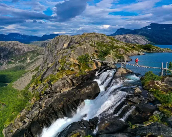En person på en hängbro över ett vattenfall under en vandringsresa i Norge. Dramatiskt landskap med berg och en blå sjö.